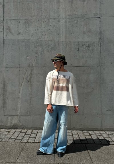 Camisa de manga larga de corte relajado en crema con estampado gráfico, combinada con jeans de pierna ancha en azul claro; accessorios incluyen un sombrero de cubo de camuflaje y gafas de sol.