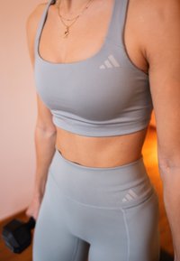 Femme portant une tenue de sport gris clair tenant un haltère noir, visible du cou aux hanches dans un environnement intérieur.