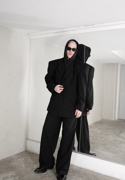 Chaqueta de traje negra de gran tamaño con grandes hombreras, sudadera con capucha negra y pantalones anchos. Se lleva con gafas de sol negras y zapatos puntiagudos.