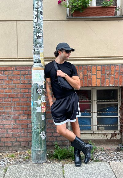 Camiseta negra, pantalones cortos deportivos negros con ribete blanco y botas negras hasta la rodilla. Apoyado contra un poste verde cubierto de graffiti.