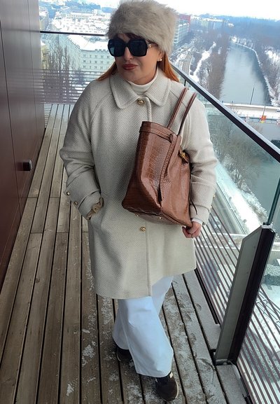 Mujer con abrigo crema, sombrero de piel, gafas de sol grandes y pantalones blancos, de pie en un balcón nevado sosteniendo un bolso marrón con textura.