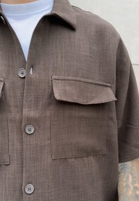 Brązowa koszula z krótkim rękawem wykonana z teksturowanej tkaniny, z dwoma kieszeniami na piersi i zapinana na guziki z przodu. Posiada szare guziki i kołnierzyk.