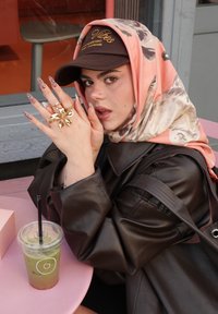 Eine Frau mit einem floral gemusterten Schal und brauner Mütze posiert mit einem goldenen Blumenring. Ein grünes Getränk mit Strohhalm steht auf einem rosa Tisch.