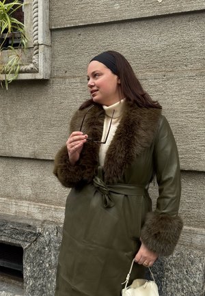 Mujer con abrigo verde de cuero con cuello y puños de piel sostiene gafas de sol y bolso blanco, parada contra una pared de piedra.