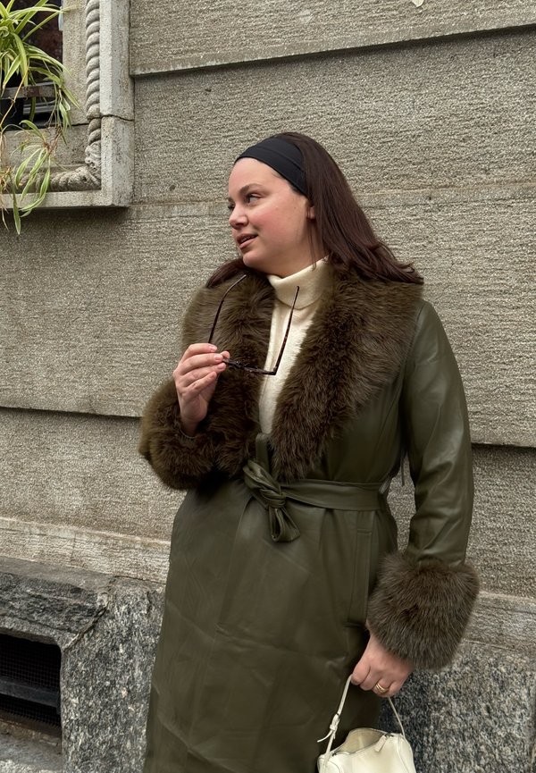 Vrouw in een groene leren jas met een bontkraag en -manchetten houdt een zonnebril en een witte handtas vast, staand tegen een stenen muur.