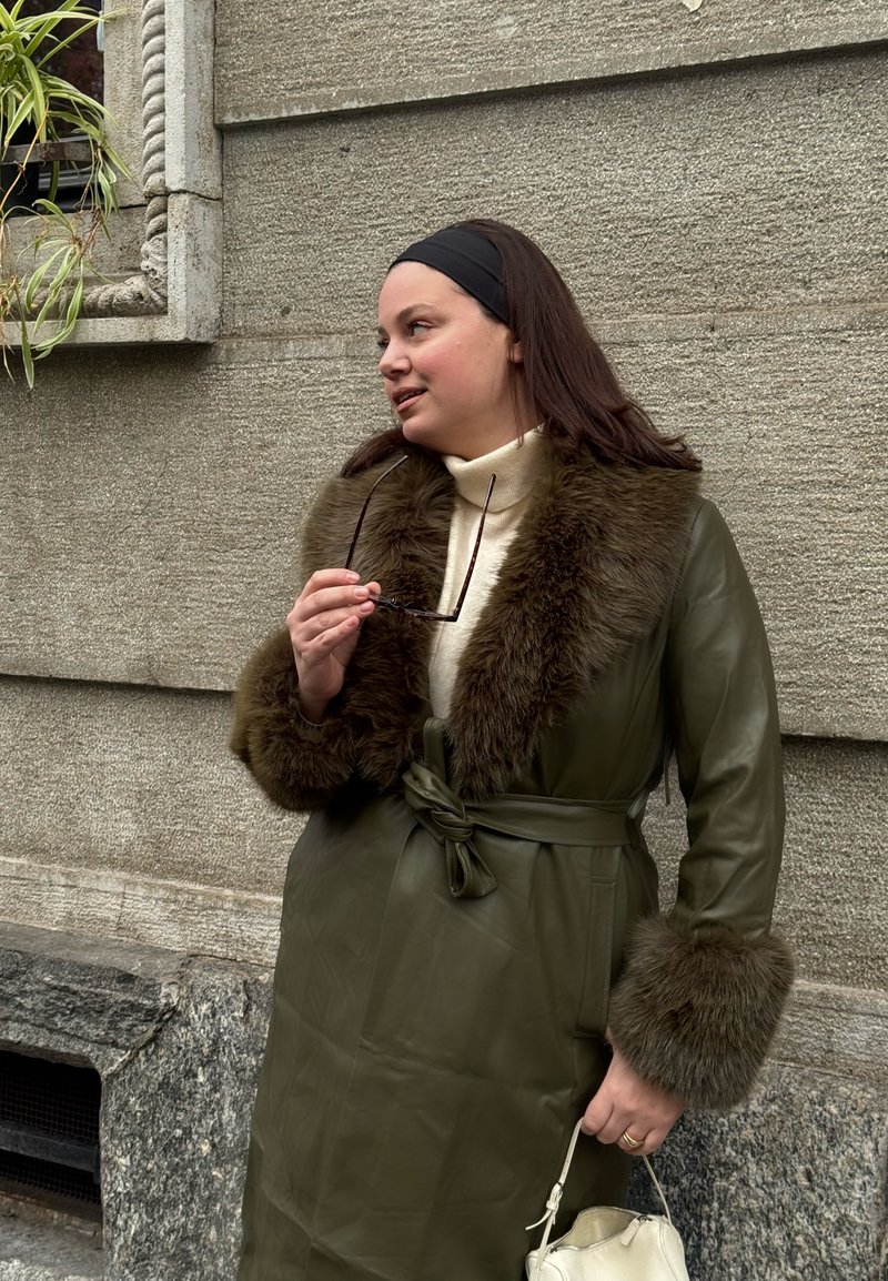 Femme vêtue d'un manteau en cuir vert avec col et poignets en fourrure, tenant des lunettes de soleil et un sac à main blanc, debout contre un mur de pierre.