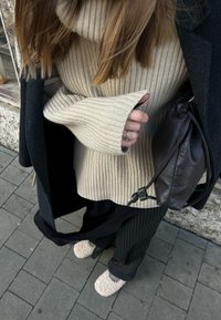 Beige bordázott garbópulóver, fekete kabát, csíkos nadrág, barna bőr táska és bolyhos bézs papucsok a járdán.