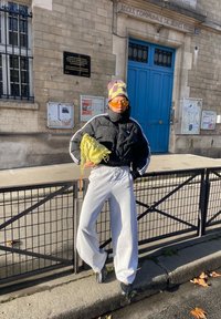 Personne portant une doudoune noire, un pantalon de survêtement gris, des lunettes de soleil jaunes et un bonnet à carreaux, tenant un sac jaune, debout devant une porte bleue.