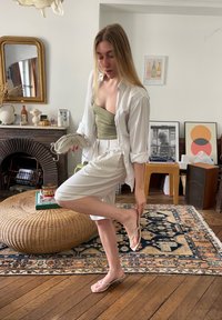 Lightweight olive green top paired with a white oversized shirt and knee-length white shorts. The outfit is completed with pale flip-flops. The setting is indoors with textured decor.