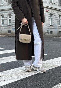Sac bandoulière beige avec ferrures dorées, manteau long brun foncé, pantalons larges blancs et baskets gris clair sur un passage piéton.