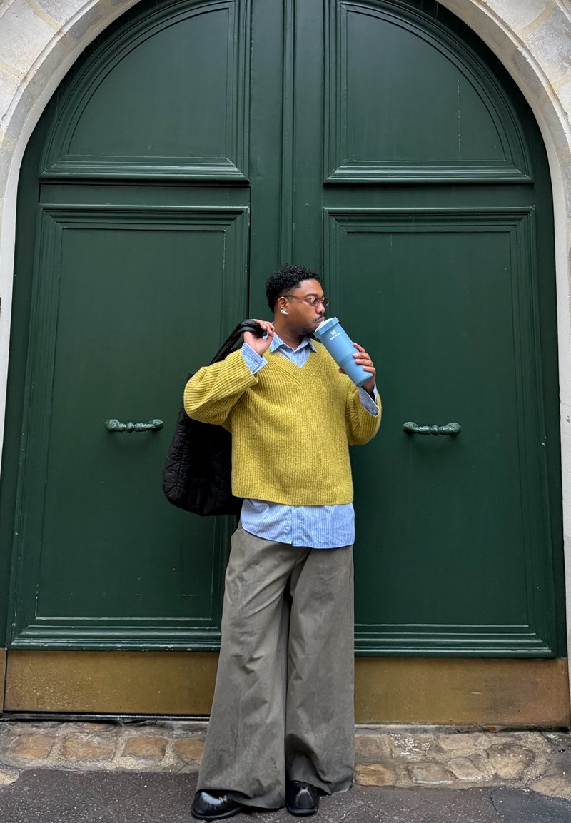 Maglione giallo a maglia sopra una camicia a righe blu, pantaloni grigi a gamba larga, con una bottiglia d'acqua blu in mano, in piedi vicino a una porta doppia verde.