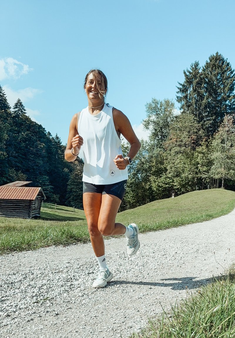 Donna che corre su un sentiero di ghiaia circondato da colline verdi e alberi sotto un cielo blu chiaro, indossando una maglietta bianca senza maniche e pantaloni corti.