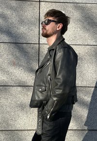 Fekete bőr motoros dzseki ezüst cipzárakkal és gombokkal, öves derékrésszel és vállpántokkal, sötét nadrággal és napszemüveggel párosítva.