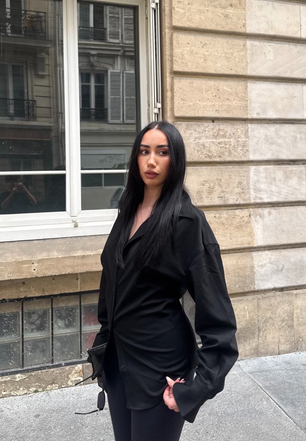 Femme aux longs cheveux noirs portant une chemise noire oversize et tenant des lunettes de soleil, debout sur un trottoir en ville près d'une fenêtre d'un bâtiment en pierre.