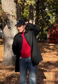 Juodas vilnonis paltas dėvimas virš raudono megztinio, derinant su mėlynomis džinsais. Fone matyti medžiai ir medinė konstrukcija.