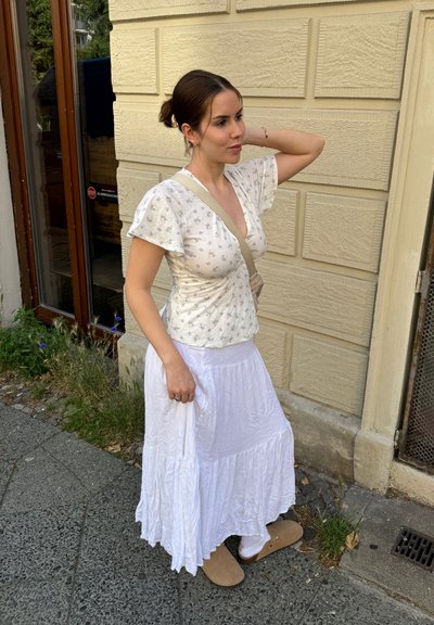 Falda blanca y ligera con capas en diferentes niveles, combinada con una blusa de estampado floral. Zapatos cómodos de tipo slip-on. Entorno natural con un camino de piedra texturizada.