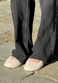 Flauschige, cremefarbene Hausschuhe mit rundem Zehendesign und einem kleinen Schleifenakzent. Getragen mit schwarzen weit geschnittenen Jeans auf einer Steinoberfläche.