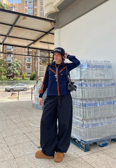 Pantalones de mezclilla oscuros de pierna ancha combinados con una chaqueta con cremallera azul marina y naranja. Calzado marrón completa el look con un fondo de botellas de agua apiladas.