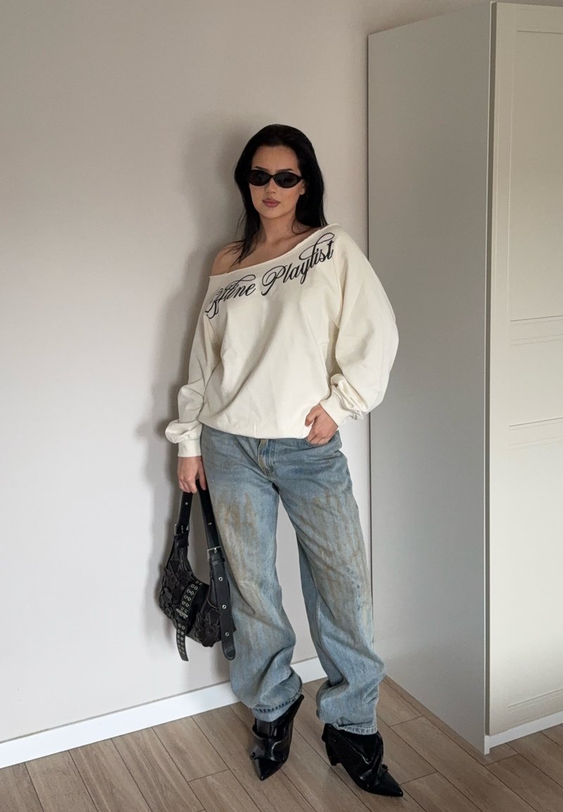Femme portant un sweat-shirt blanc à épaules dénudées avec l'inscription "Offline Playlist", un jean large délavé, des bottes noires pointues, des lunettes de soleil, tenant un sac à main noir à l'intérieur.