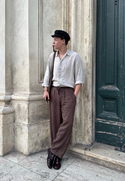Camisa de lino de botones en gris claro, pantalones de lino marrón, gorra negra y zapatos de cuero marrón. De pie frente a una pared de piedra texturizada.