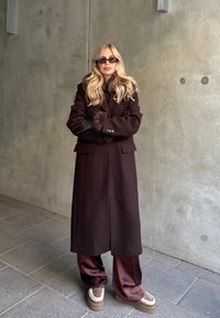 Manteau en laine marron avec poignets en fourrure, superposé à une tenue en satin marron. Porte des lunettes de soleil foncées et des chaussures épaisses avec des accents crème. Fond neutre.