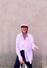 Ροζ ριγέ πουκάμισο με κουμπιά, ανοιχτό μωβ φούτερ από κάτω και ένα ταιριαστό ροζ καπέλο με λουλουδάτο λογότυπο. Μαύρο παντελόνι και λευκή τσάντα.