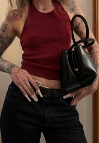 Haut débardeur rouge sans manches, jean noir taille haute, et un petit sac à main noir. Ongles longs et pointus remarquables et tatouages visibles sur les bras.