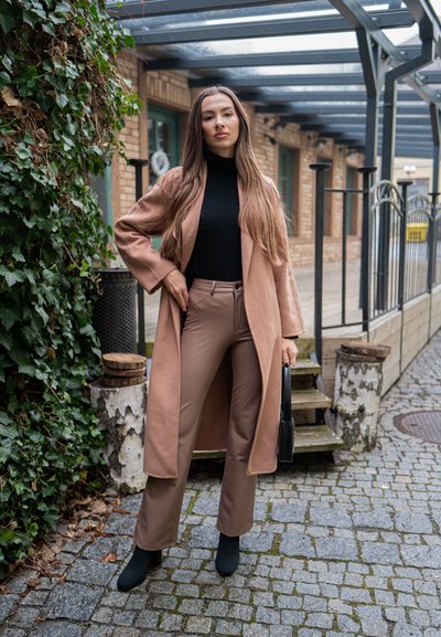 Abrigo largo marrón sobre un suéter negro de cuello alto, combinado con pantalones marrones y botas negras hasta el tobillo. El escenario incluye pavimento de piedra y hiedra.