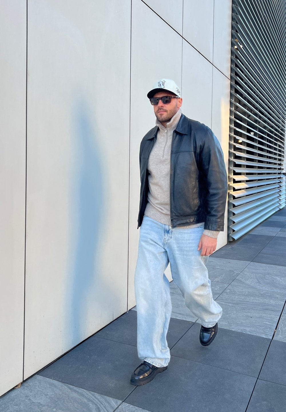 Μαύρο δερμάτινο μπουφάν πάνω από ένα ανοιχτό γκρι πουλόβερ με ψηλό λαιμό, μπλε τζιν και μαύρα παπούτσια. Αξεσουάρ περιλαμβάνουν ένα λευκό καπέλο και γυαλιά ηλίου.