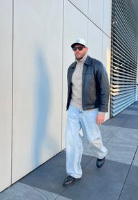 Μαύρο δερμάτινο μπουφάν πάνω από ένα ανοιχτό γκρι πουλόβερ με ψηλό λαιμό, μπλε τζιν και μαύρα παπούτσια. Αξεσουάρ περιλαμβάνουν ένα λευκό καπέλο και γυαλιά ηλίου.