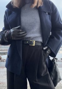 Osoba nosząca czarne skórzane rękawiczki, granatowy płaszcz, szary sweter, czarne spodnie z skórzanym paskiem oraz niosąca czarną skórzaną torbę na zewnątrz.