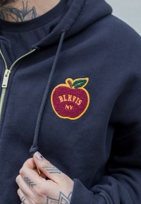 Položena navy bluza s kapuco, ki se zapenja na zadrgo, z teksturirano vezenino rdeče jabolka v rumenih in zelenih odtenkih. Vključuje vrvico in kovinsko zadrgo.