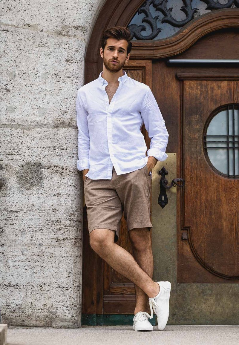 Witte button-up shirt, bruine shorts en witte sneakers. Model leunt tegen een gestructureerde stenen muur, met een houten deur met een ronde raam.