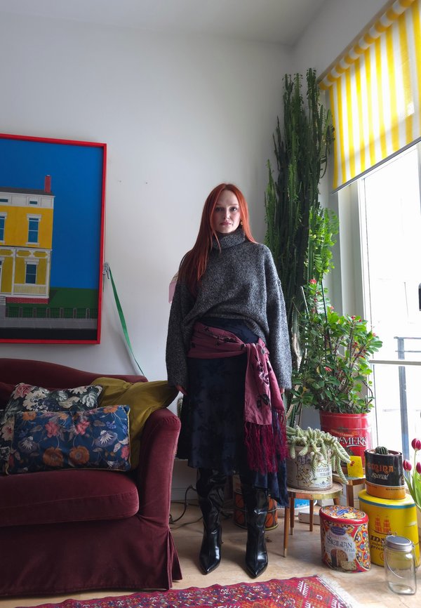 Donna con capelli rossi che indossa un maglione grigio, una gonna scura e una sciarpa, in piedi accanto a un divano rosso e piante vicino a una finestra con una tenda a righe gialle.