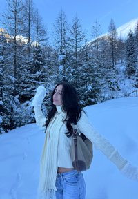 Femme en pull blanc, écharpe et moufles, debout les bras écartés dans une forêt enneigée, portant un sac à bandoulière beige et marron.