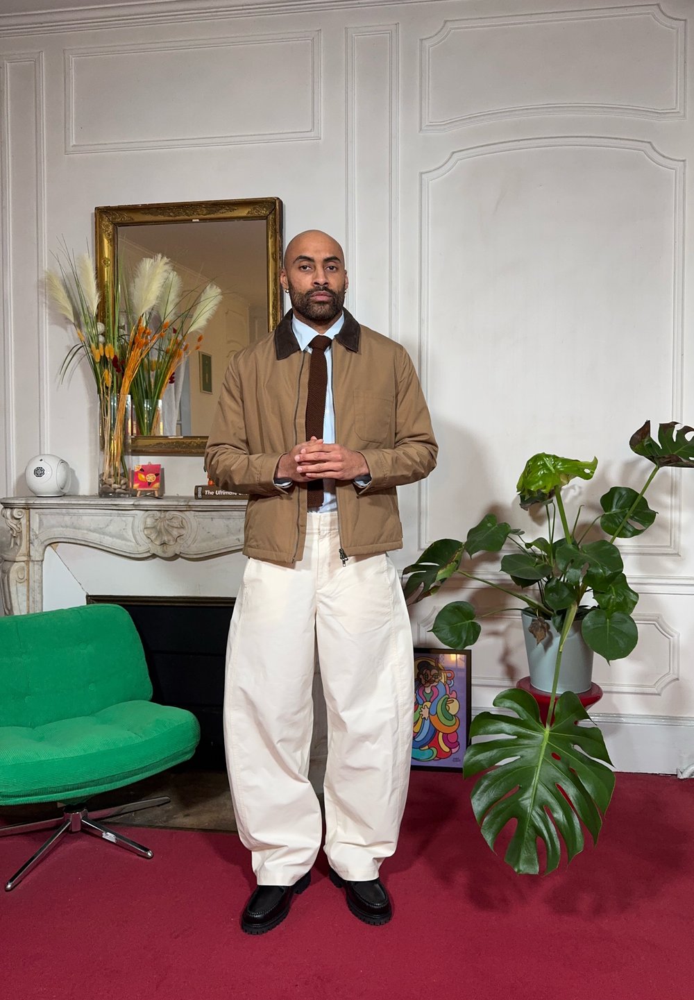 Veste marron à fermeture éclair avec un col contrastant, chemise bleu clair, cravate foncée et pantalon blanc à jambes larges. Chaussures noires. Chaise verte et plante en arrière-plan.