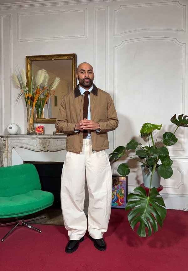 Veste marron à fermeture éclair avec un col contrastant, chemise bleu clair, cravate foncée, et pantalon blanc à jambes larges. Chaussures noires. Chaise verte et plante en arrière-plan.