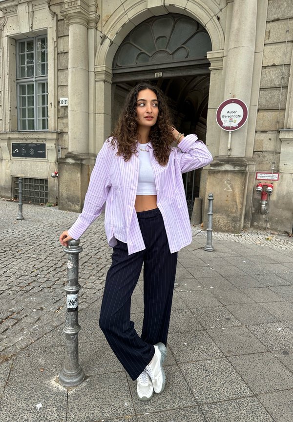 Una camicia oversize a righe lilla su un crop top bianco e pantaloni a righe blu scuro, abbinata a sneaker grigie e bianche.