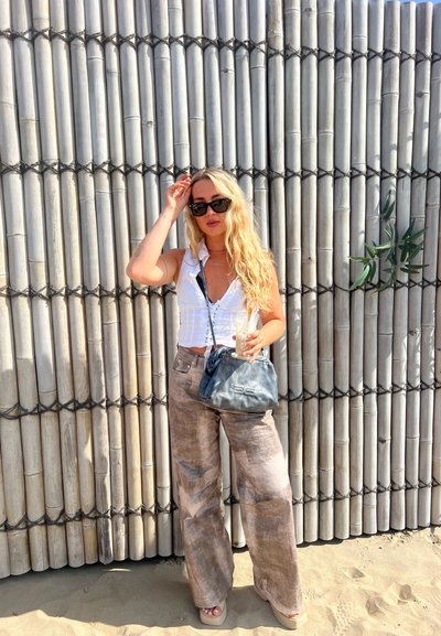 Top corto blanco ligero, pantalones de pierna ancha marrón con estampado, bolso gris, gafas de sol negras. Fondo de bambú y suelo de arena visibles.