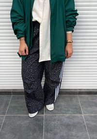 Casaco verde extragrande por cima de uma camisa branca com botões, combinado com calças largas pretas com padrão de leopardo e riscas laterais brancas. Sapatos brancos.