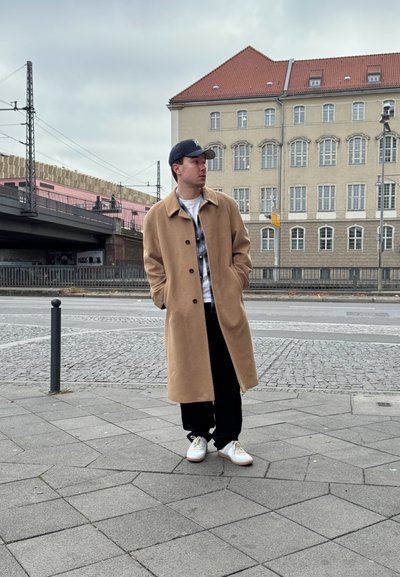 Abrigo de lana beige con botones en el frente, usado sobre una camisa de cuadros y una camiseta blanca. Pantalones negros y zapatillas blancas completan el atuendo.