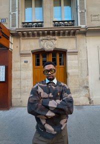 Homme portant de grandes lunettes carrées et un pull camouflage, debout les bras croisés devant une porte en bois ornée et un bâtiment en pierre beige.