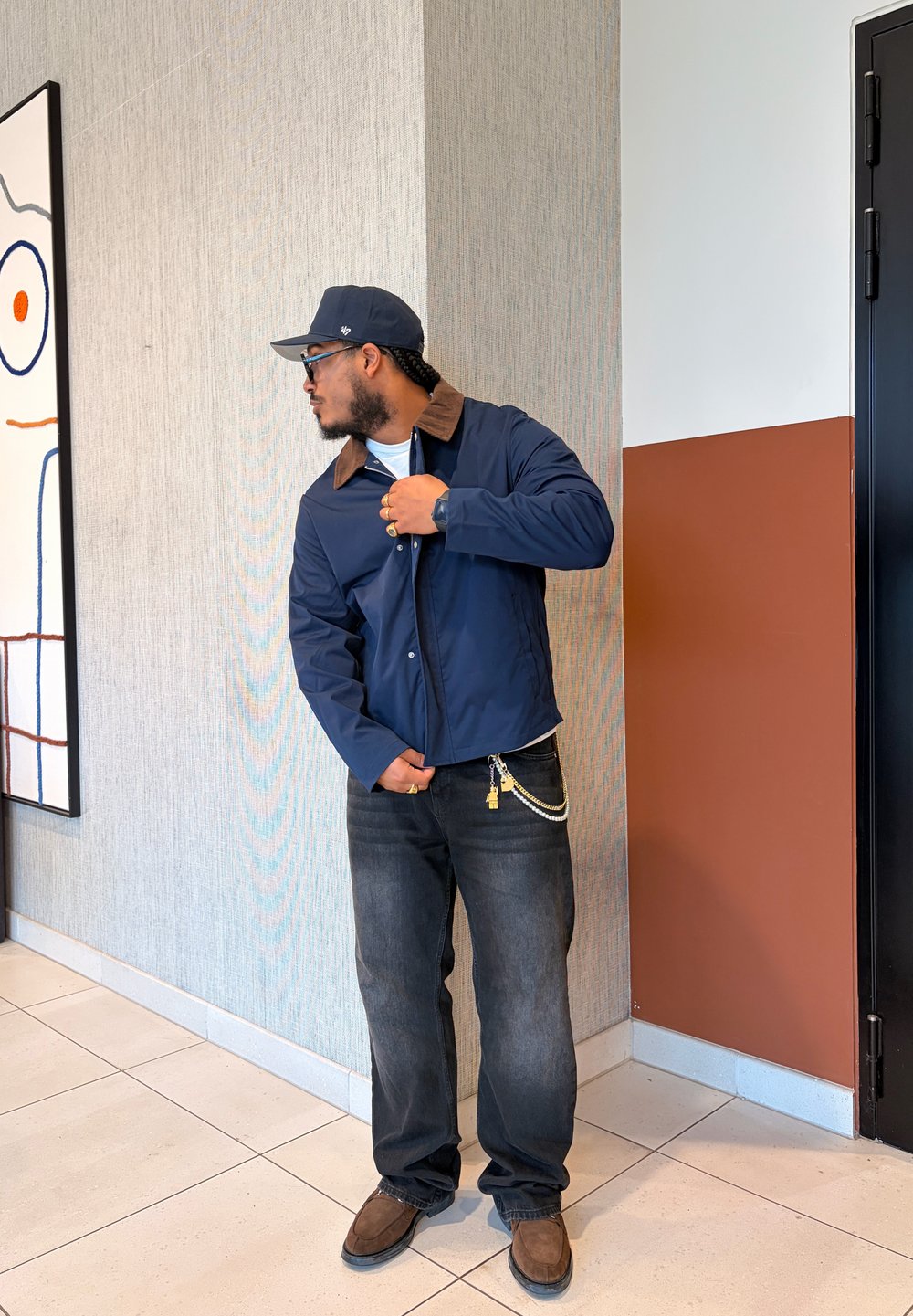 Veste marine avec un col marron, associée à un jean foncé. Accessoires comprenant une chaîne et une montre. En arrière-plan, une œuvre d'art géométrique.
