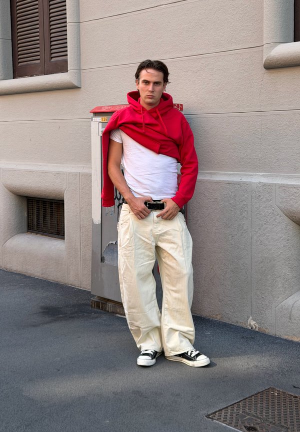 Joven con camiseta blanca y sudadera roja sobre los hombros, pantalones beige y zapatillas negras, de pie junto a una pared beige cerca de una caja de suministros.