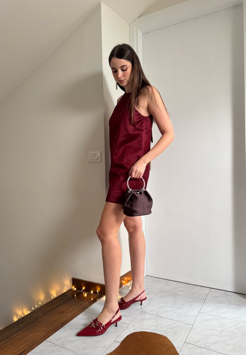 Femme en robe bordeaux sans manches et talons assortis, tenant un petit sac à main rond, debout à l'intérieur près d'une porte blanche et de guirlandes lumineuses.