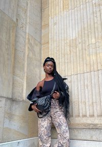 Zwarte leren jas, zwarte tanktop, camouflage-patroon broek, zilveren ringoorbellen, staande tegen gestructureerde stenen kolommen.