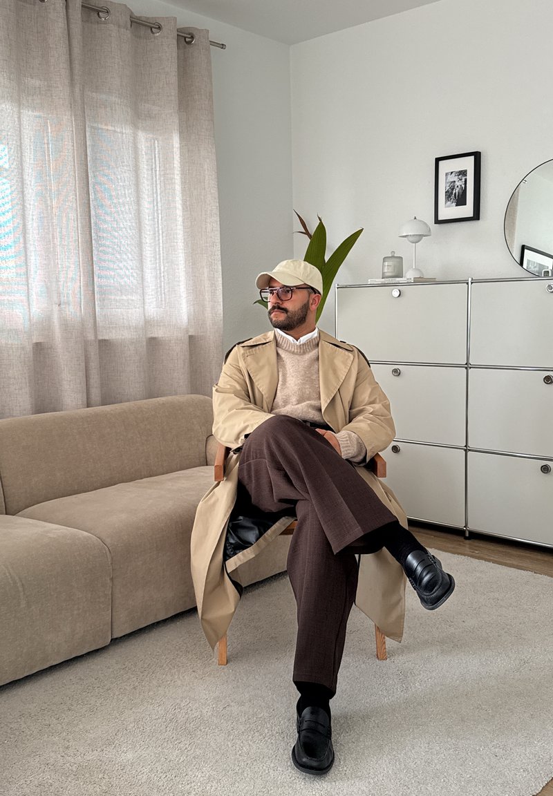 Mand iført beige trenchcoat, hat og briller, der sidder korslagte ben på en træstol i et minimalistisk stue med sofa og skab.