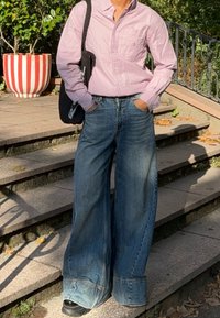 Person iført en rosa stripete skjorte og vide blå jeans som står på utendørs steintrapper med hendene i lommene og en svart skulderveske.