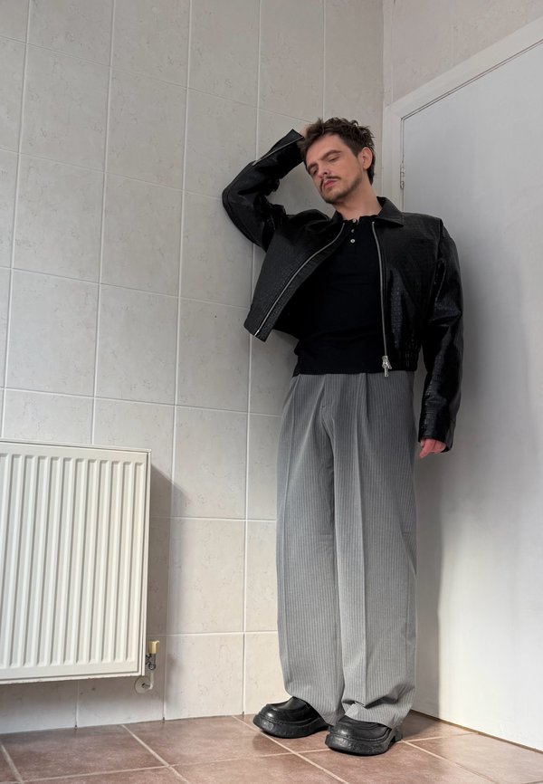 Hombre con los ojos cerrados, vestido con chaqueta negra y pantalones grises anchos, de pie junto a una pared embaldosada cerca de una puerta blanca cerrada y un radiador.