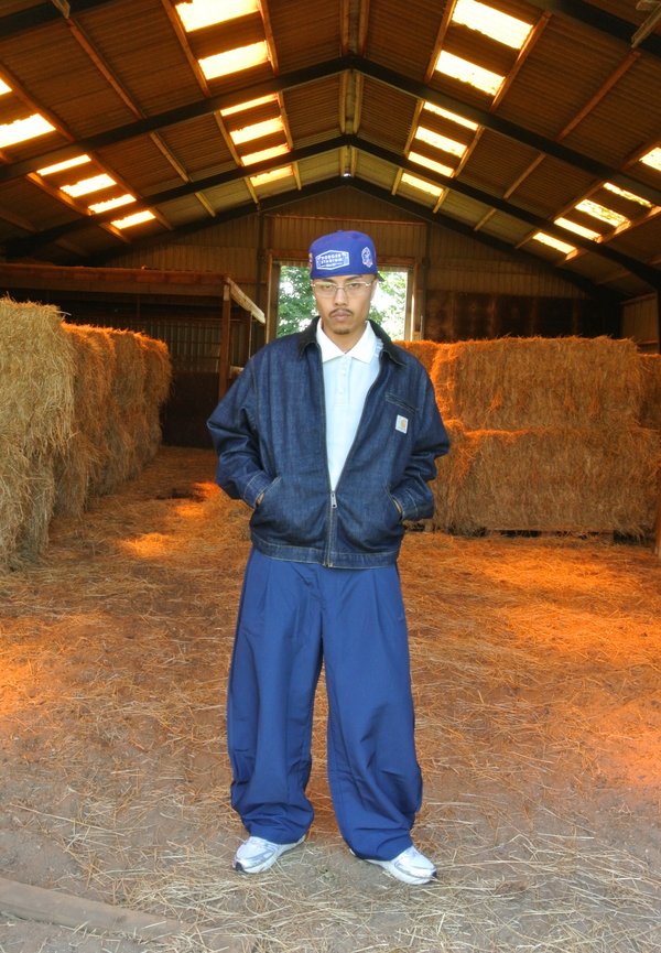Homme portant une veste bleue, une casquette et un pantalon large debout dans une grange avec des bottes de foin empilées le long des murs et la lumière du soleil venant des fenêtres du toit.